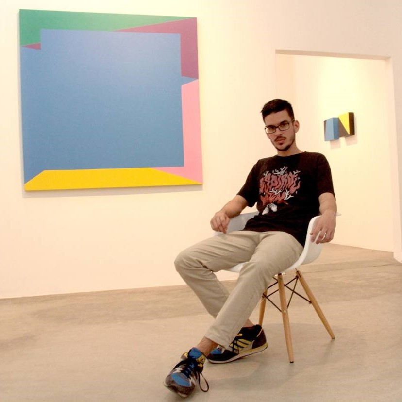 Javier Vivas en su exposición posando sentado al lado de una de sus obras